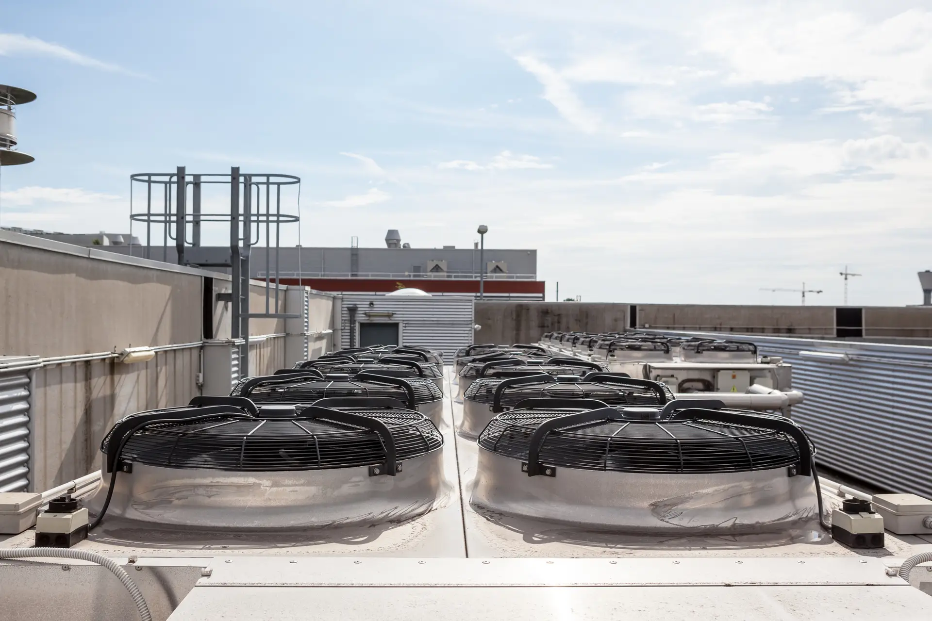 Rows of industrial HVAC units are mounted on a rooftop under a partly cloudy sky. The metal units, part of commercial air conditioning repair efforts, have visible fans and are surrounded by various pipes and structures. Buildings and construction cranes can be seen in the background.