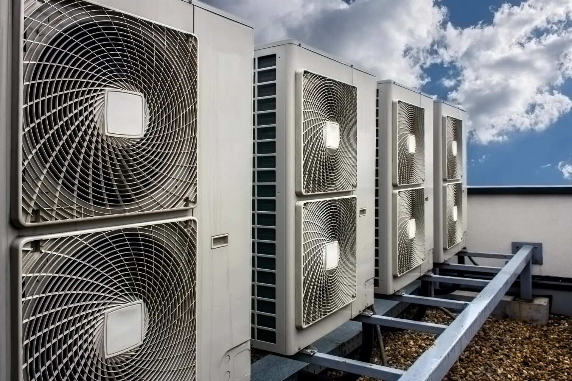 A row of five outdoor air conditioning units is neatly perched on a rooftop metal framework. These commercial HVAC units, with their visible fan grilles and metallic casings, stand aligned under a partly cloudy sky, providing an industrial charm against the backdrop of blue and white.