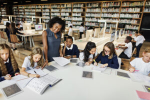 A teacher assists a diverse group of students working on assignments at a table in a library equipped with bookshelves, computers, and an advanced Building Automation System in the background.