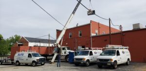 A crane lifts equipment onto the roof of a red brick building, possibly installing a Building Automation System, as three white HVAC service vans are parked nearby and a worker directs the operation.