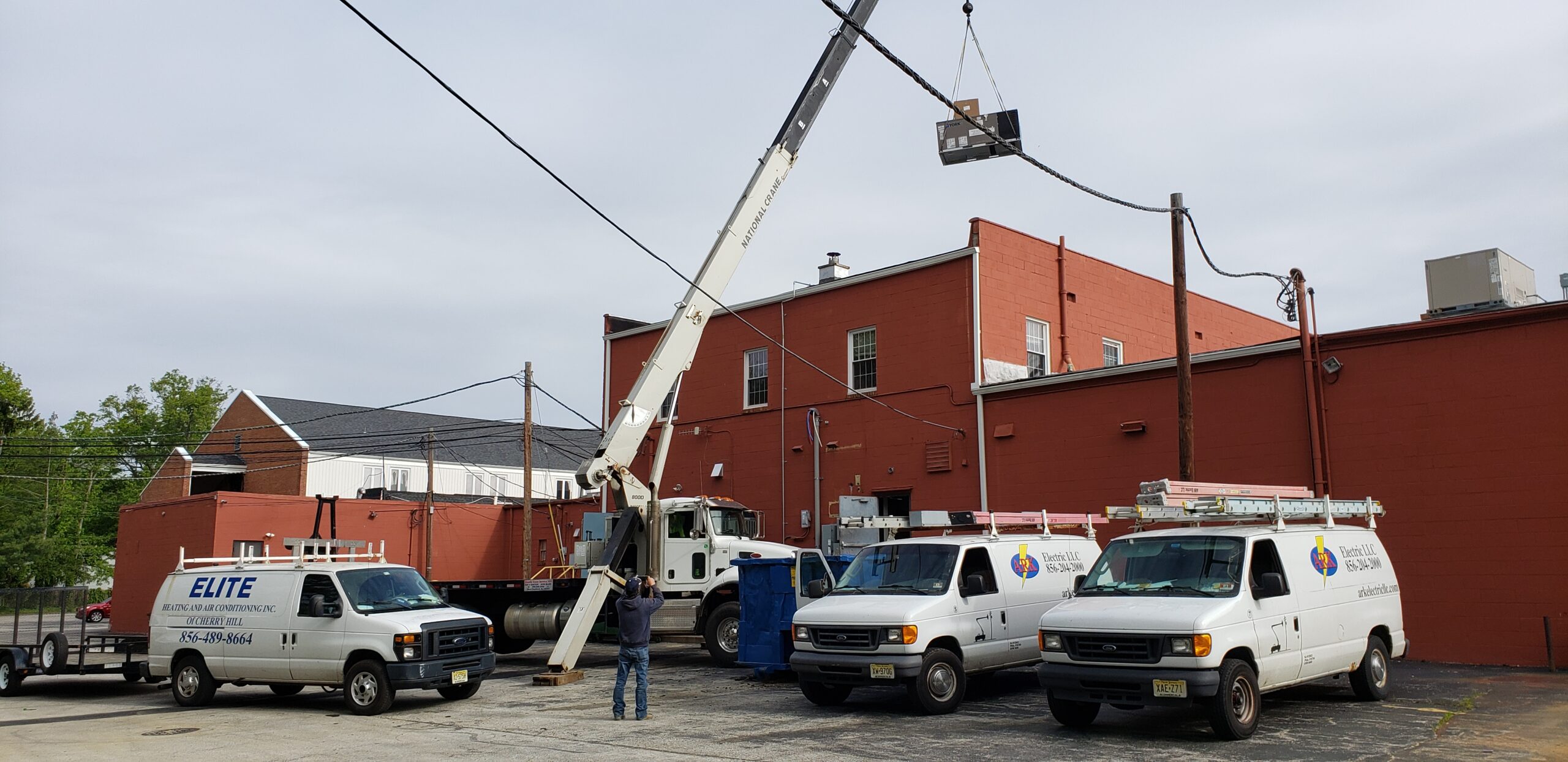 A crane lifts equipment onto the roof of a red brick building, possibly installing a Building Automation System, as three white HVAC service vans are parked nearby and a worker directs the operation.