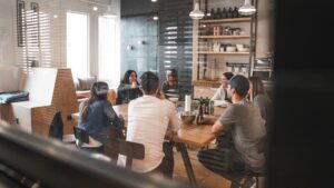 A group of people sit around a wooden table having a meeting in a modern, well-lit office space equipped with a Building Automation System, with shelves and kitchen items in the background.