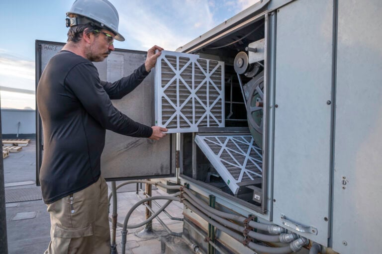 A worker wearing a hard hat and safety glasses replaces air filters in an outdoor HVAC unit on a rooftop, ensuring the Building Automation System runs efficiently.