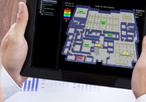 A person holds a tablet displaying a digital floor plan with labeled rooms and color-coded indicators, showcasing features of a Building Automation System.