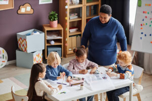 Four children sit at a table drawing with markers while an adult stands nearby in a classroom, which is equipped with shelves, boxes, a whiteboard, and an advanced Building Automation System ensuring comfort.