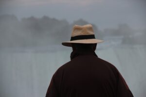 A person wearing a tan hat and dark coat stands with their back to the camera, quietly observing a misty waterfall beyond—nature’s flow in contrast to the precision of a Building Automation System.