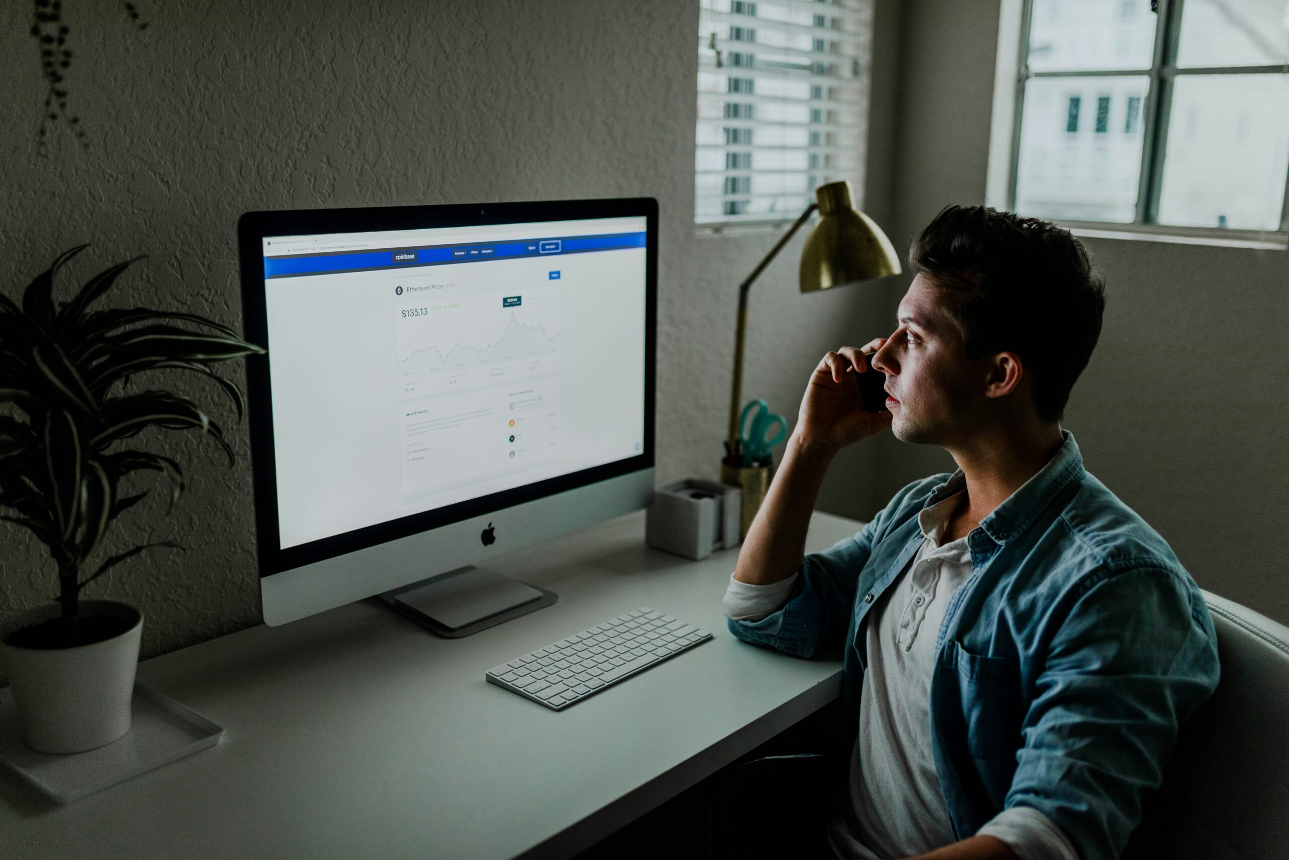 A man sits at a desk in his home office, looking at a computer screen displaying Building Automation System graphs while talking on the phone.