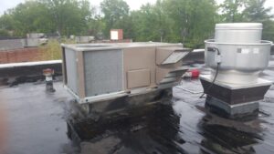 Rooftop HVAC unit and ventilation fan installed on a flat, black rooftop with trees and buildings visible in the background, seamlessly integrated into the Building Automation System for efficient climate control.