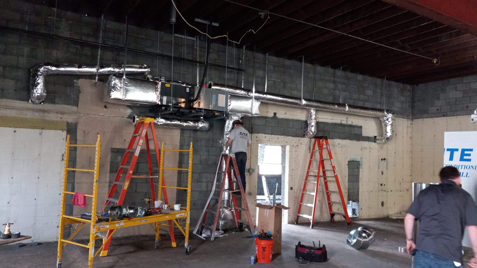 Workers install ductwork and HVAC components on a wall inside a building under construction, preparing the space for integration with the Building Automation System; ladders and tools are visible around the workspace.