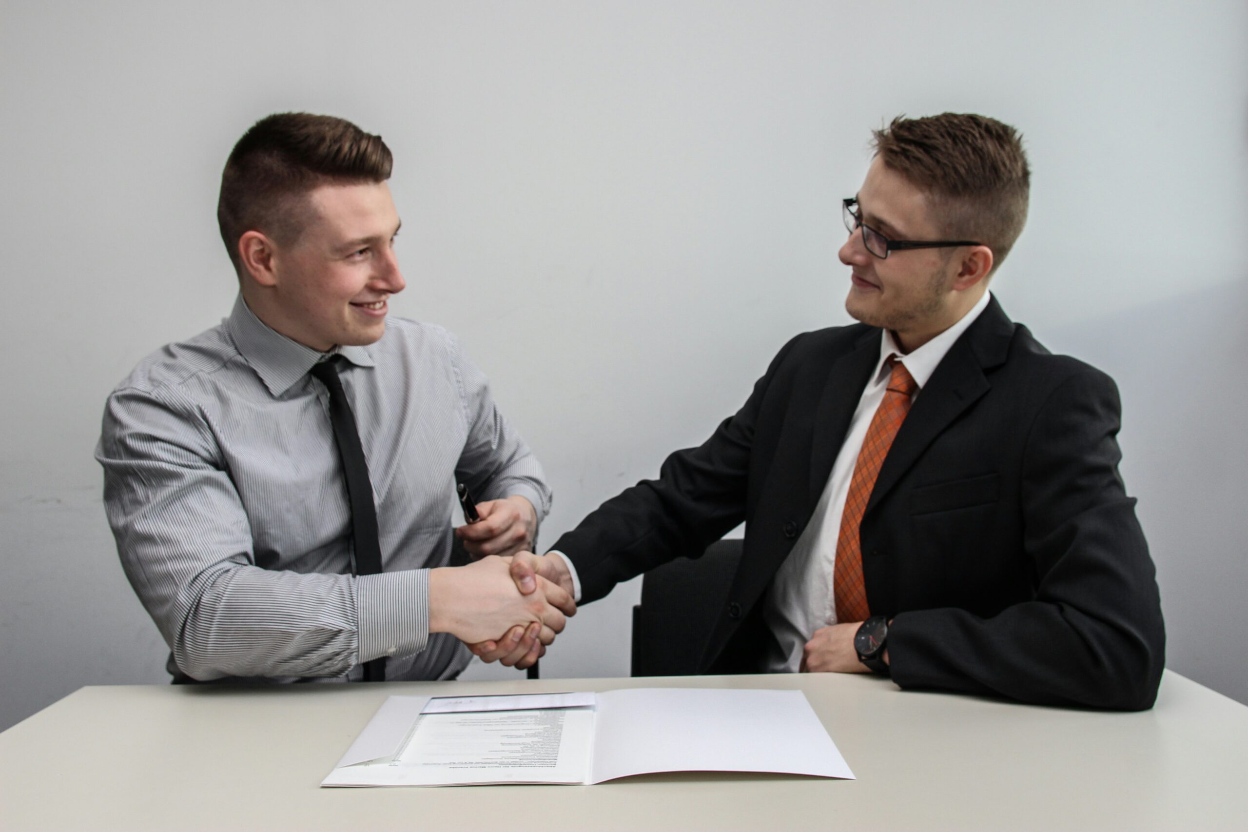 Two men in business attire shake hands across a table with documents, indicating a successful meeting or agreement on implementing a Building Automation System.