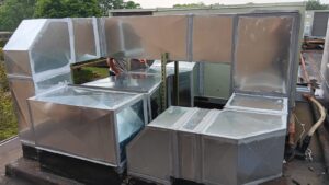 Large metal HVAC ductwork installation on a rooftop, with a worker partially visible behind the ducts. The setup is designed for seamless integration with a Building Automation System; trees are visible in the background.
