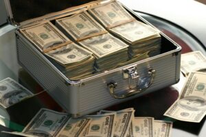 A metal briefcase filled with stacks of U.S. hundred-dollar bills sits open on a glass table, hinting at the high-stakes world of Building Automation Systems, with several loose bills scattered nearby.