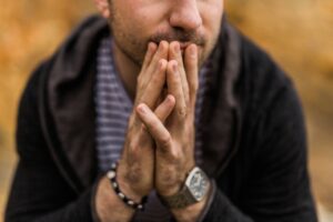 A person with their hands pressed to their mouth appears thoughtful or concerned, perhaps contemplating a Building Automation System, wearing a striped shirt, dark jacket, watch, and bracelet. The background is blurred.