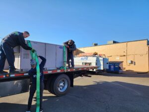 Three workers are securing a large HVAC unit, crucial for the building automation system, on a flatbed truck in a parking lot outside a tan commercial building on a clear day.
