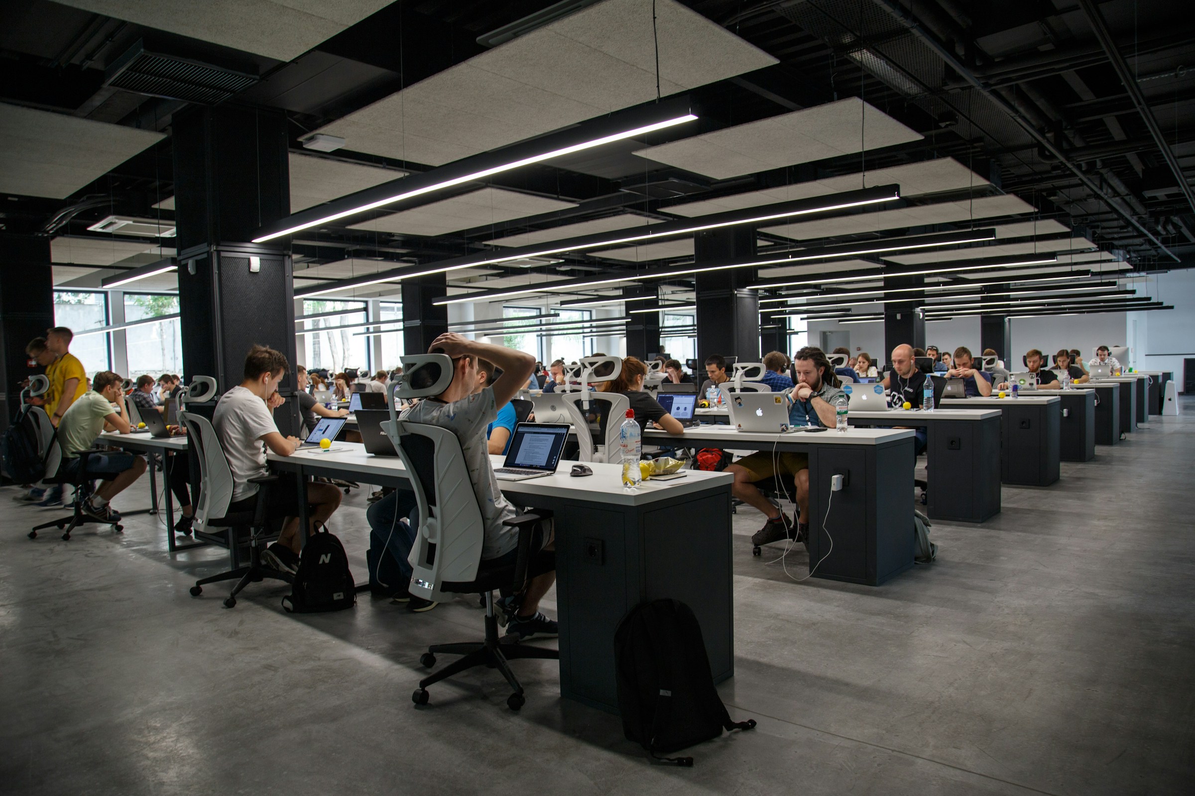 Large open-plan office with many people working at rows of desks with computers, under bright overhead lighting managed by a Building Automation System in a modern, industrial-style workspace.