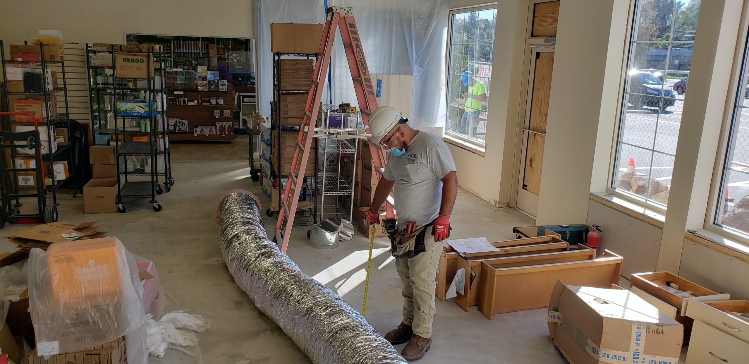 A construction worker wearing a hard hat stands inside a partially renovated room with tools, boxes, and building materials scattered around, preparing the site for the installation of a Building Automation System.
