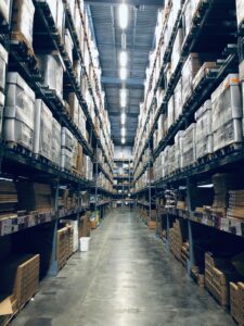 Wide aisle in a warehouse lined with tall shelves filled with stacked boxes and pallets under bright overhead lighting, efficiently managed by a Building Automation System.
