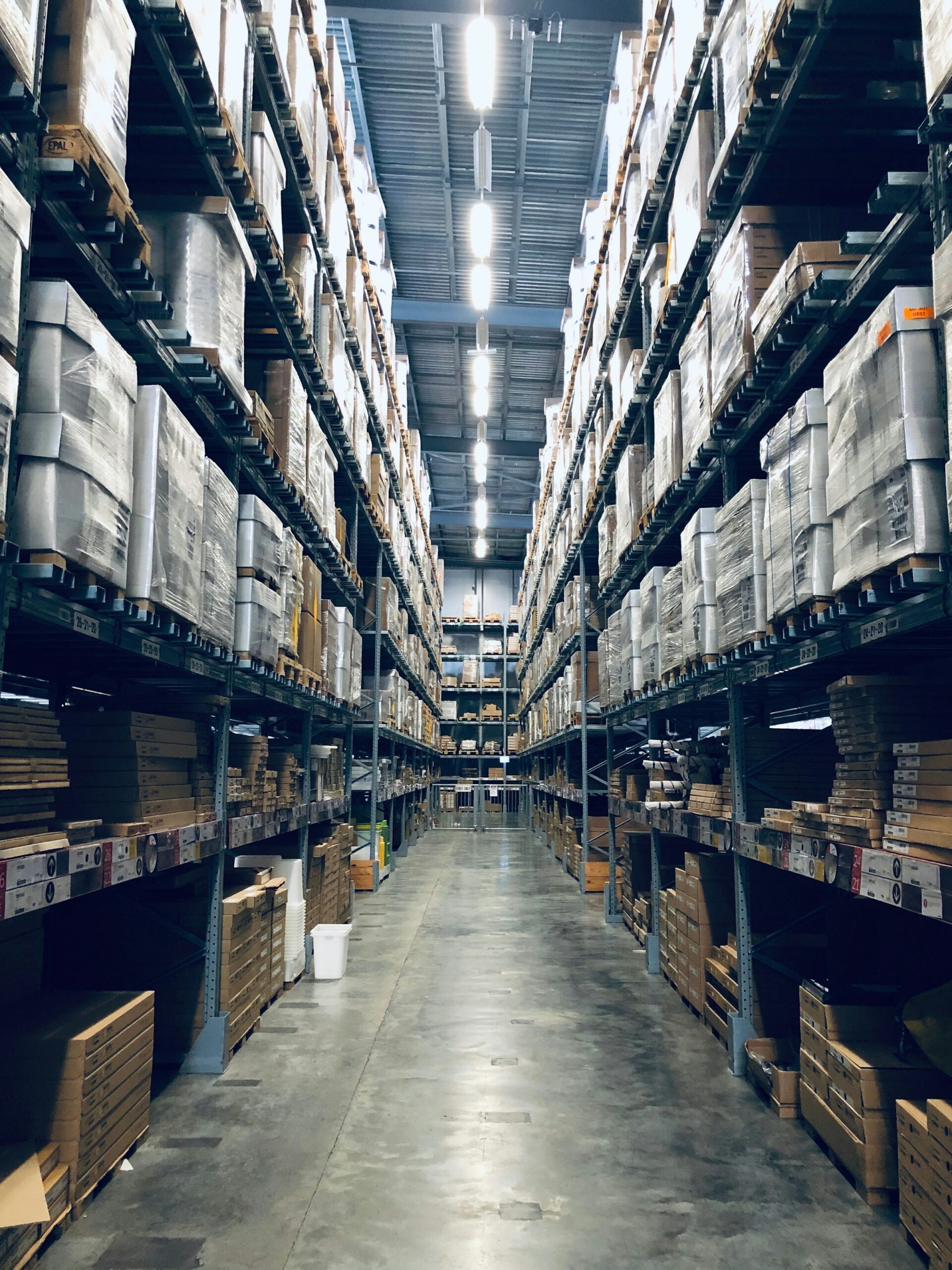 Wide aisle in a warehouse lined with tall shelves filled with stacked boxes and pallets under bright overhead lighting, efficiently managed by a Building Automation System.