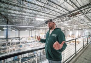 A man in a green hoodie and cap stands on a mezzanine, gesturing with his hands as he overlooks a large industrial warehouse equipped with a Building Automation System managing the equipment and materials below.