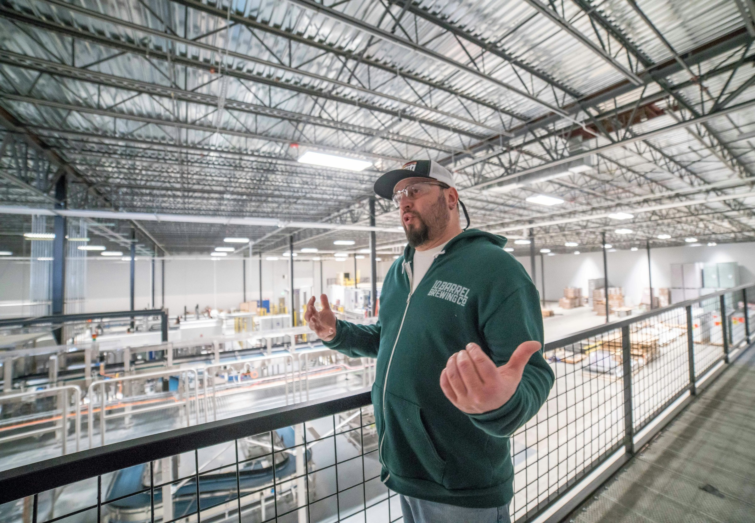 A man in a green hoodie and cap stands on a mezzanine, gesturing with his hands as he overlooks a large industrial warehouse equipped with a Building Automation System managing the equipment and materials below.