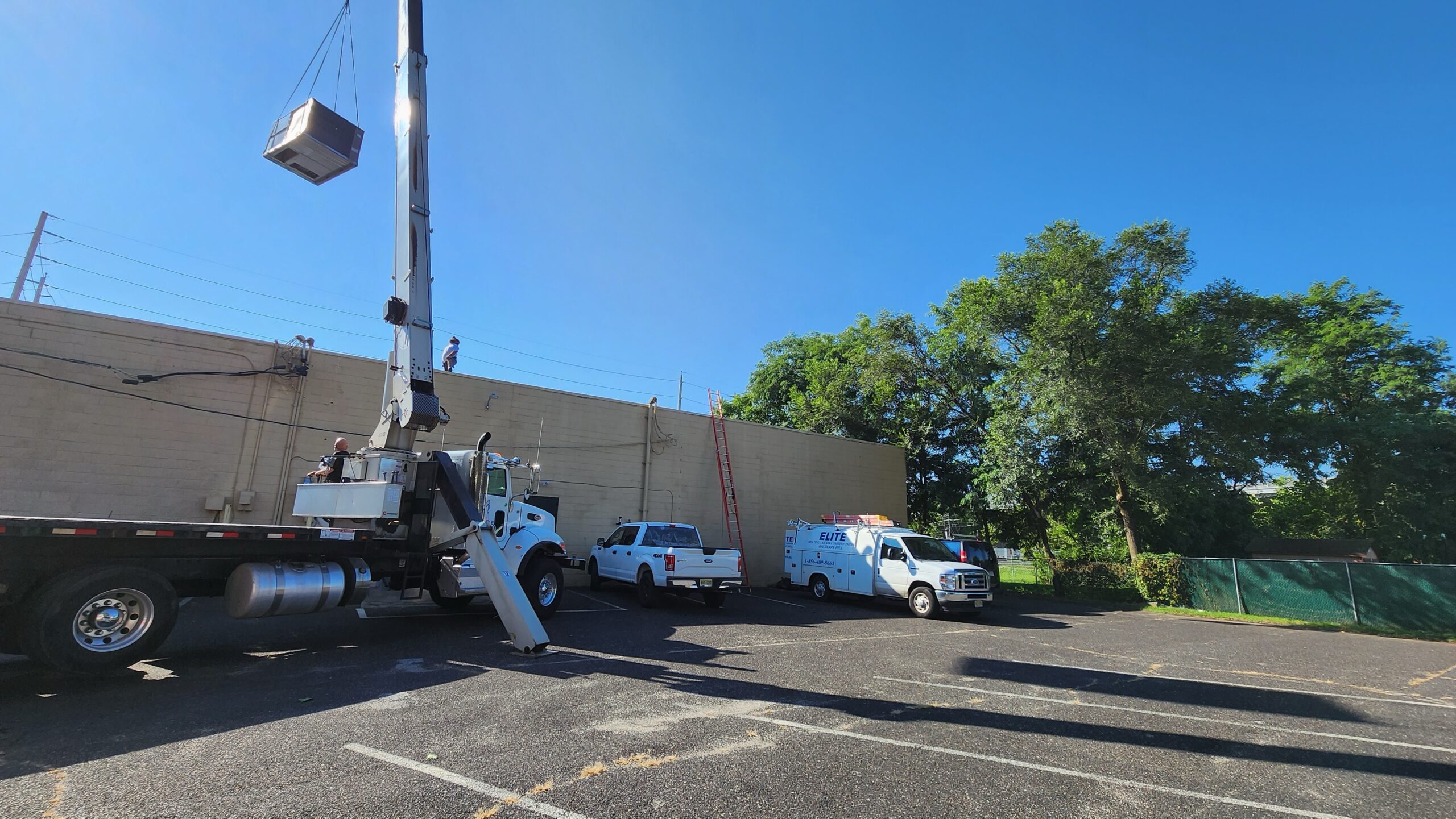 A crane lifts equipment—possibly for a Building Automation System—onto the roof of a beige building as workers and utility vans are present in a sunny parking lot.
