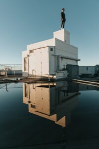 A person stands on top of a white rooftop structure, managed by a Building Automation System, reflected in a large puddle of water under a clear blue sky.