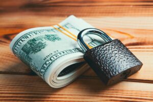 A roll of U.S. hundred dollar bills secured with a rubber band and locked with a brown padlock, placed on a wooden surface—symbolizing the security essential in any Building Automation System.