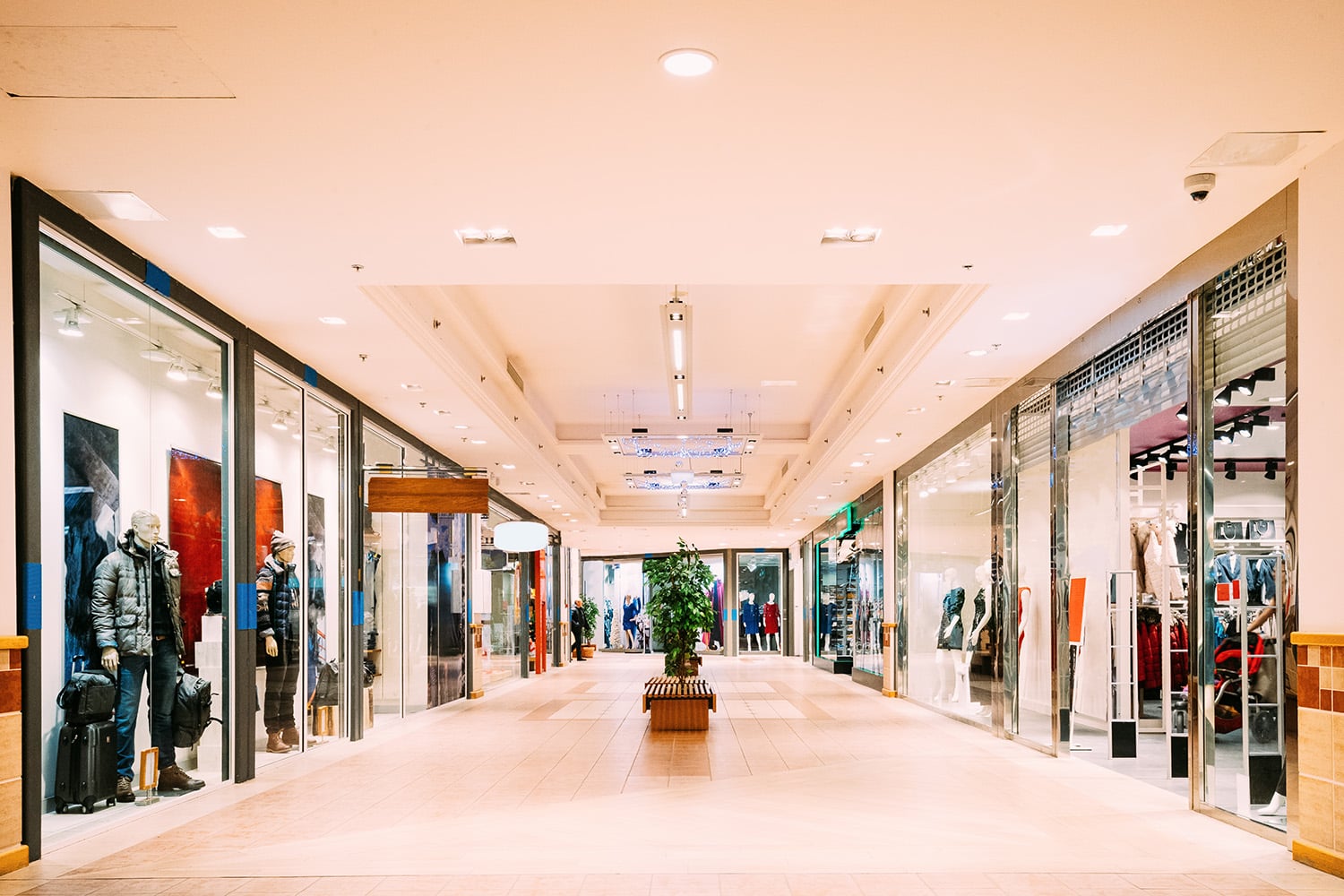 A brightly lit shopping mall corridor features storefronts on both sides, mannequins in display windows, and a bench with a potted plant in the center—all efficiently managed by a Building Automation System for optimal comfort.