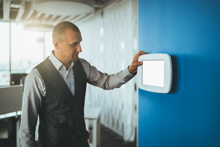 A man in business attire adjusts a wall-mounted touchscreen control panel, seamlessly managing the Building Automation System in a modern office setting.
