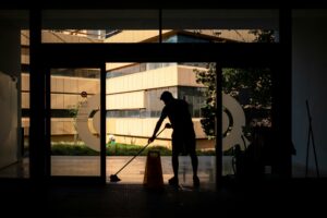A person mops the floor near a large glass entrance, with a caution sign in front. Modern buildings and an advanced Building Automation System are visible outside.