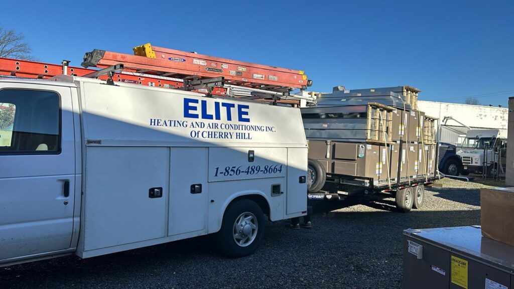 A white ELITE Heating and Air Conditioning van with ladders on top is parked next to a trailer loaded with HVAC units in a gravel lot, ready for Building Automation System installation.