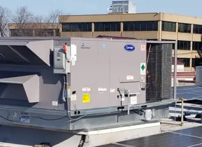 On a clear day, a rooftop HVAC unit with gray panels, a red switch, and metal grating sits against the skyline. The unit's array of stickers highlights its upkeep by top commercial air conditioning repair professionals. In the background, other buildings and lush trees complete the scene.