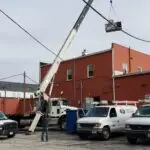 A crane lifts an HVAC unit onto the roof of a red brick building. Below, three white service vans with ladders and the Elite brand, known for commercial HVAC repair, are parked. Two workers observe the operation, with one guiding the crane. Power lines run overhead and a tree is visible in the background.