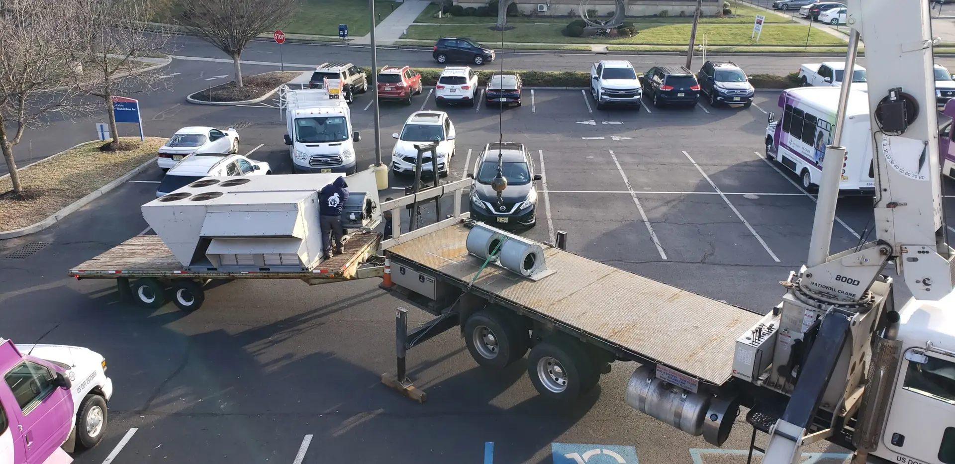 A flatbed truck loaded with large HVAC units is parked in a lot where a worker from one of the best HVAC companies near you is adjusting the units. Nearby are several cars and a delivery truck. In the background, a purple and white bus stands beside a leafless tree and building under the clear sky.