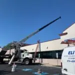 A crane truck with a long arm is stationed by Burlington Coat Factory for Air Conditioning repair. An Elite Heating and Air Conditioning van is parked in front, while a ladder leans against the building wall. Two parking spaces with blue lines are visible, one marked by a cone and a meter.
