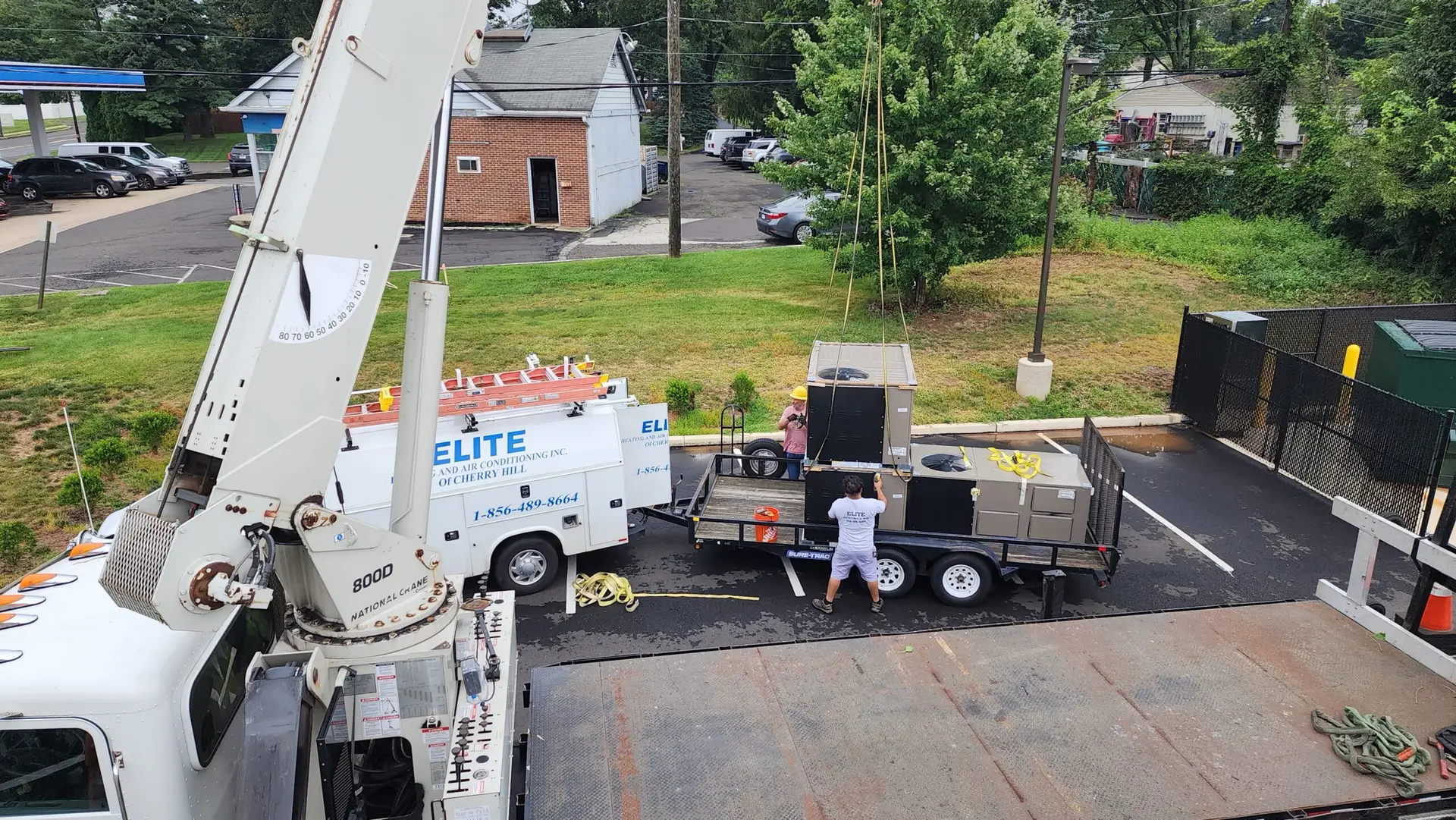 A crane lifts HVAC equipment from a flatbed trailer beside a white Elite-branded utility truck in a parking lot. Two workers guide the equipment, demonstrating excellence in commercial air conditioning repair. Trees, buildings, and a street form the backdrop.