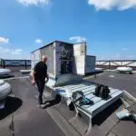 A person stands on a rooftop next to an open HVAC unit, which reveals visible mechanical components. Tools and a backpack rest on the ground nearby. Several other closed units are on the flat roof, bordered by a wooden railing. The sky is clear with scattered clouds.
