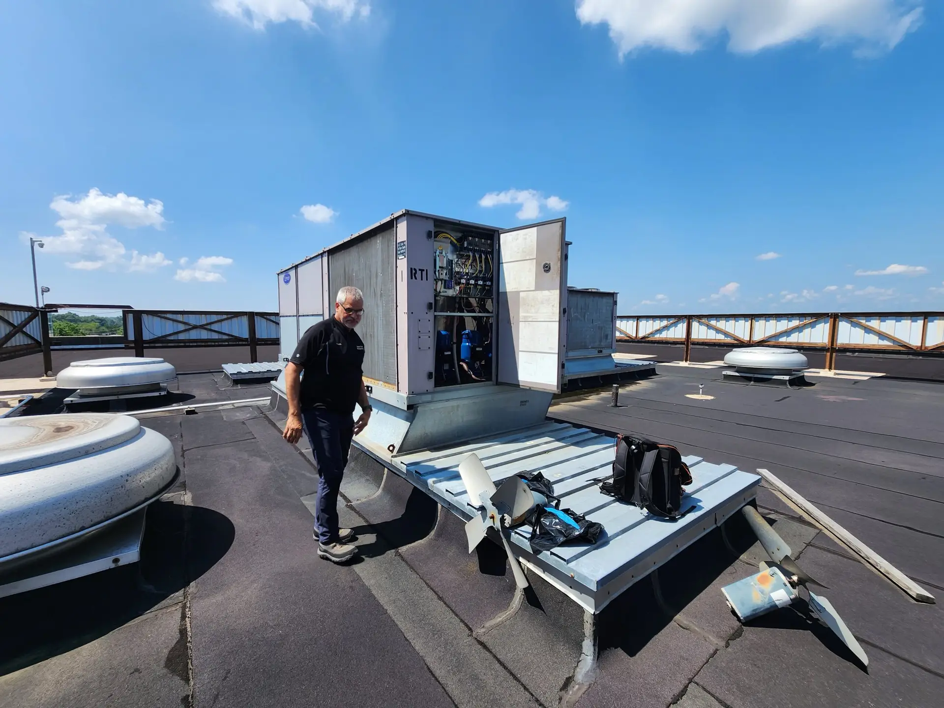 A person stands on a rooftop next to an open HVAC unit, which reveals visible mechanical components. Tools and a backpack rest on the ground nearby. Several other closed units are on the flat roof, bordered by a wooden railing. The sky is clear with scattered clouds.