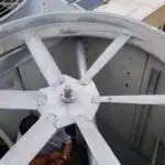 A person conducting an hvac inspection inside a large, industrial cooling fan housing on a New Jersey rooftop. The metallic fan's long blades surround them as they adjust components. The scene reveals rooftop elements like vents, a smaller fan, and a side view of the building.