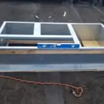 A rectangular metal frame sits on a flat, black rooftop. An orange bucket is on the left, with an orange extension cord leading to a power tool from one of the best HVAC companies near me on the right. A blue level rests across the frame, surrounded by small wooden boards and tools.