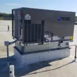 A commercial air conditioning unit from York rests on a flat roof under a clear blue sky. The unit, perfect for repair needs near me, has ventilated sides and is mounted on a metal platform. Nearby, various pipes and conduit lines weave through the scene, with trees lining the horizon in the background.