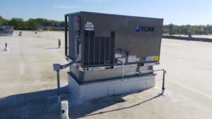 A commercial air conditioning unit from York rests on a flat roof under a clear blue sky. The unit, perfect for repair needs near me, has ventilated sides and is mounted on a metal platform. Nearby, various pipes and conduit lines weave through the scene, with trees lining the horizon in the background.