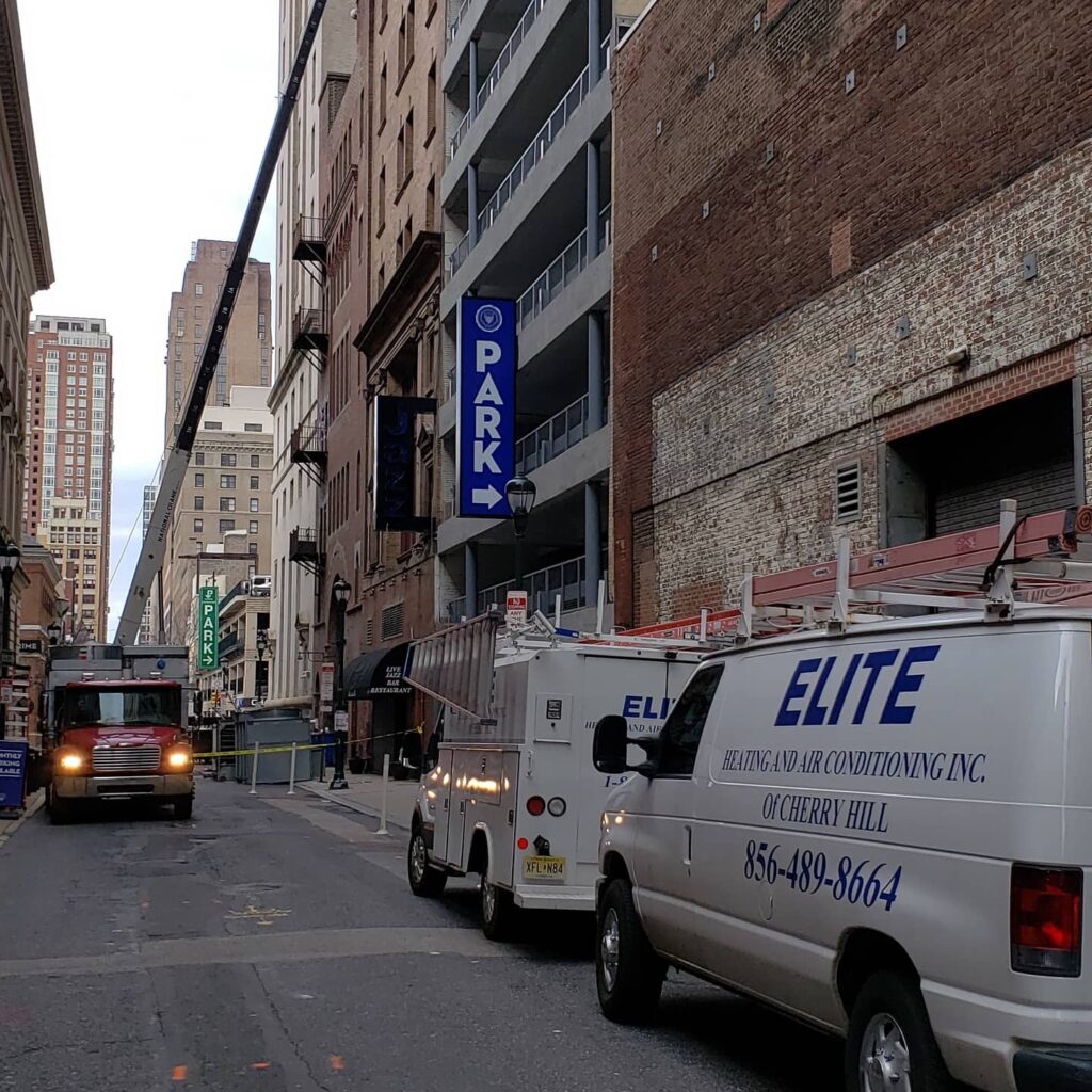 A narrow city street with service vehicles parked along the right side, including Elite Heating and Air Conditioning vans that rank among the best air conditioning repair companies near me. A fire truck is in the distance. Tall buildings line the street, and a sign indicates Park with an arrow towards a nearby parking facility.