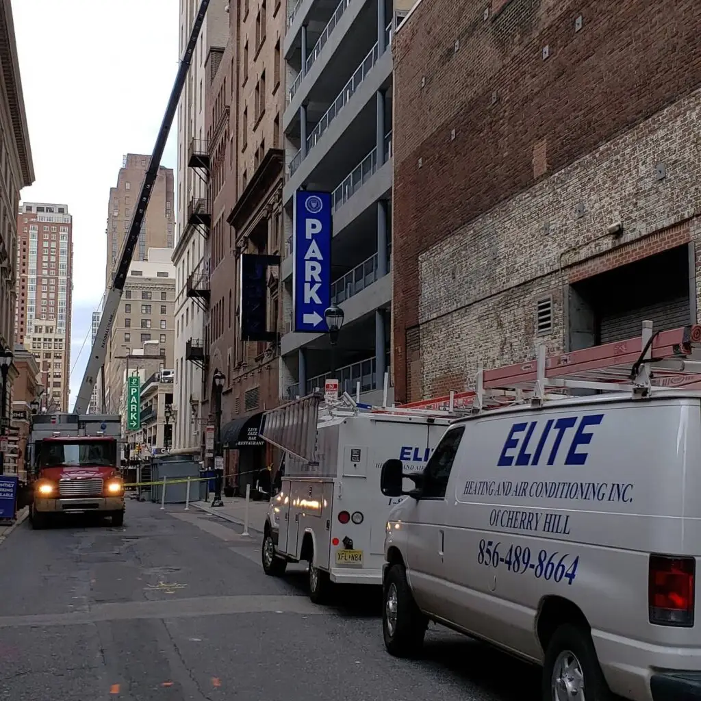 A narrow city street with service vehicles parked along the right side, including Elite Heating and Air Conditioning vans that rank among the best air conditioning repair companies near me. A fire truck is in the distance. Tall buildings line the street, and a sign indicates Park with an arrow towards a nearby parking facility.