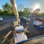 A view from above shows a bustling construction site with a central white crane truck, supported by stabilizers. Nearby, two vans labeled Elite hint at one of the best HVAC companies near me. Various vehicles and equipment are scattered around, while trees and a bright sun enhance the backdrop.