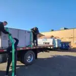 Three workers on a flatbed truck secure a large HVAC unit using green straps under a clear blue sky. One worker stands on the truck, another on the ground adjusts the strap, and the third oversees the process. A dumpster and other equipment are in the background near a beige building.