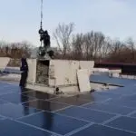 Two workers are on a flat rooftop engaged in commercial HVAC repair. One stands atop a large HVAC unit, handling a crane cable, while the other supervises. The roof is dotted with solar panels, and trees adorn the horizon under a clear sky.