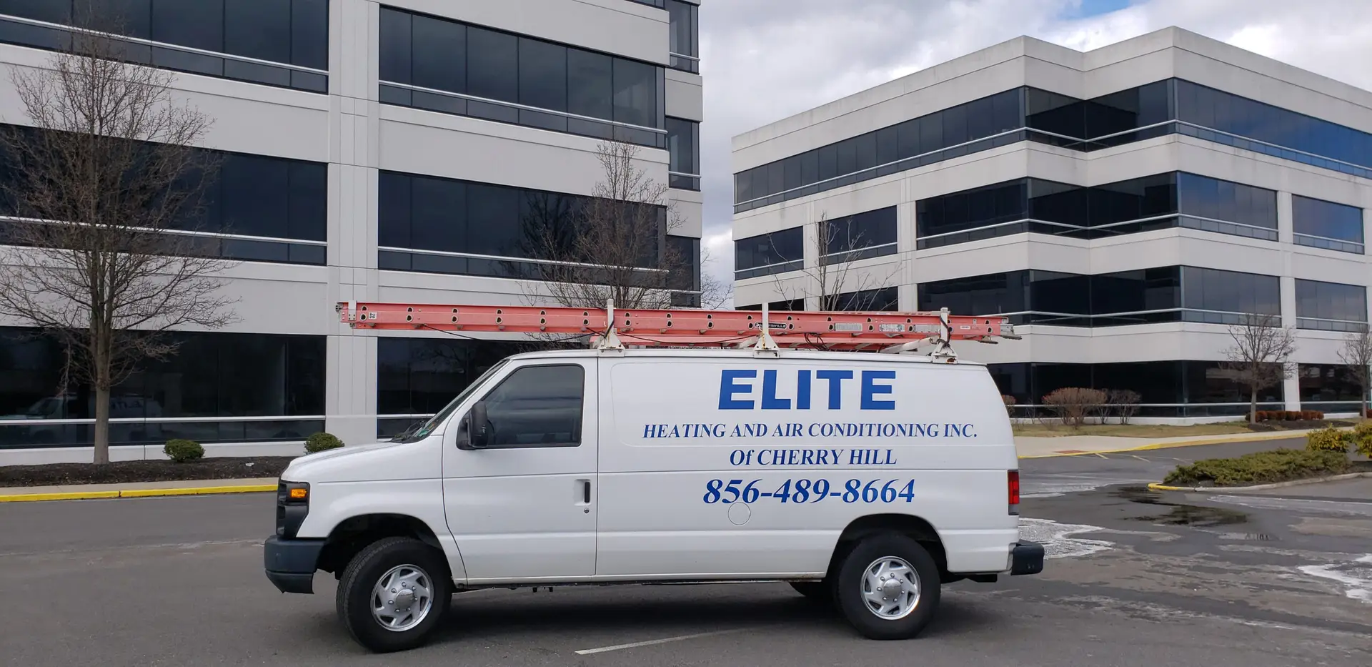 A white van with Elite Heating and Air Conditioning Inc. of Cherry Hill and a phone number is parked in front of modern office buildings. A red ladder is secured to the roof, ready for commercial HVAC repair. The sky is partly cloudy, with leafless trees lining the paved driveway.