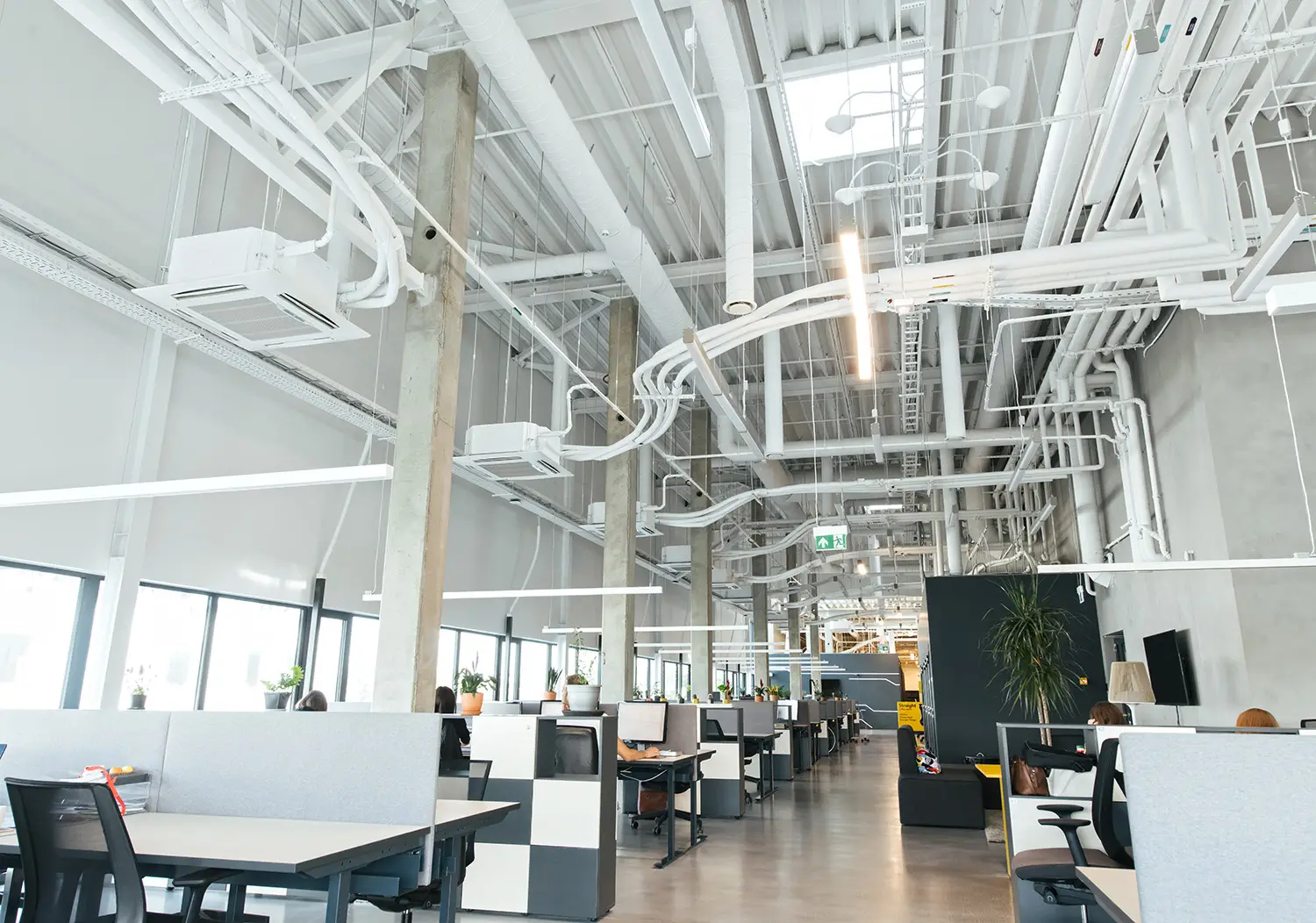 A spacious, modern open-plan office with high ceilings, exposed ductwork, and efficient commercial HVAC repair ensures comfort. Desks are arranged in rows with ergonomic chairs and computers. Large windows let in natural light while hanging lights illuminate the workspace. Some employees are working at their desks.