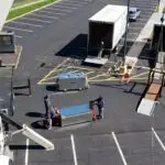 Aerial view of a parking lot bustling with activity from one of the best HVAC companies near me. Two trucks with open trailers are onsite, and several workers, including two men maneuvering a large metal crate while another operates the crane. Equipment and tools are scattered around, with cars parked in the distance.